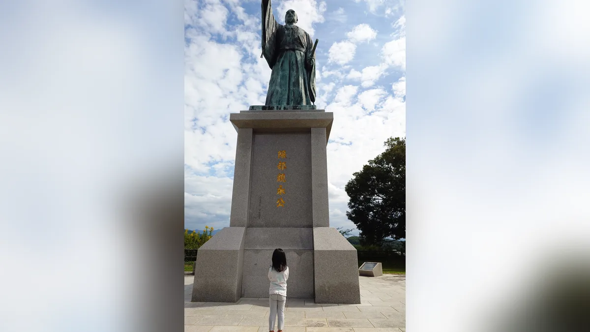 あんずの丘の人工芝すべりの上に立つ隈部親永像と、その前に立つ4歳の後ろ姿